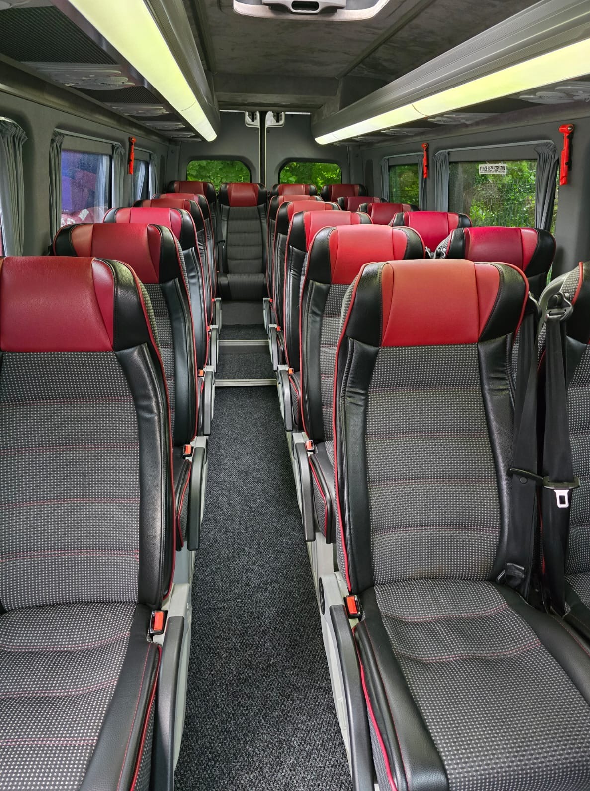 Wnętrze komfortowego busa z rzędami foteli w kolorze czarno-czerwonym. Widok na przejście i okna. Oświetlenie sufitowe.