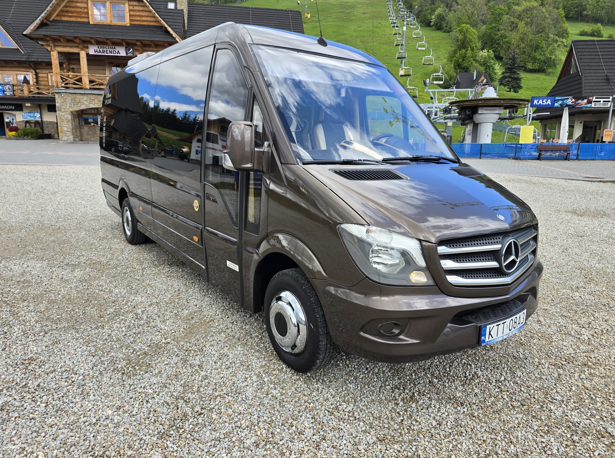 Brązowy bus Mercedes-Benz Sprinter na parkingu, z budynkiem w stylu góralskim i wyciągiem narciarskim w tle, sugerujący transport do atrakcji turystycznej.