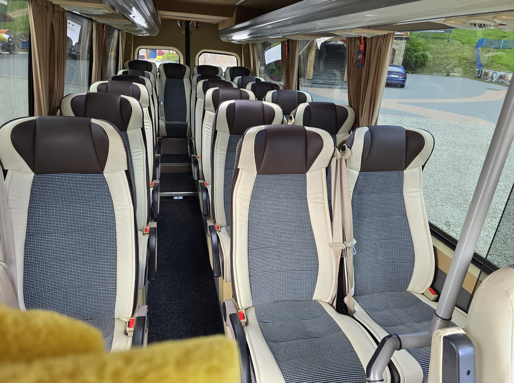 Wnętrze autobusu z rzędami foteli w beżowo-brązowej kolorystyce z pasami bezpieczeństwa. Widok na okna i otoczenie na zewnątrz. Komfortowe podróżowanie.