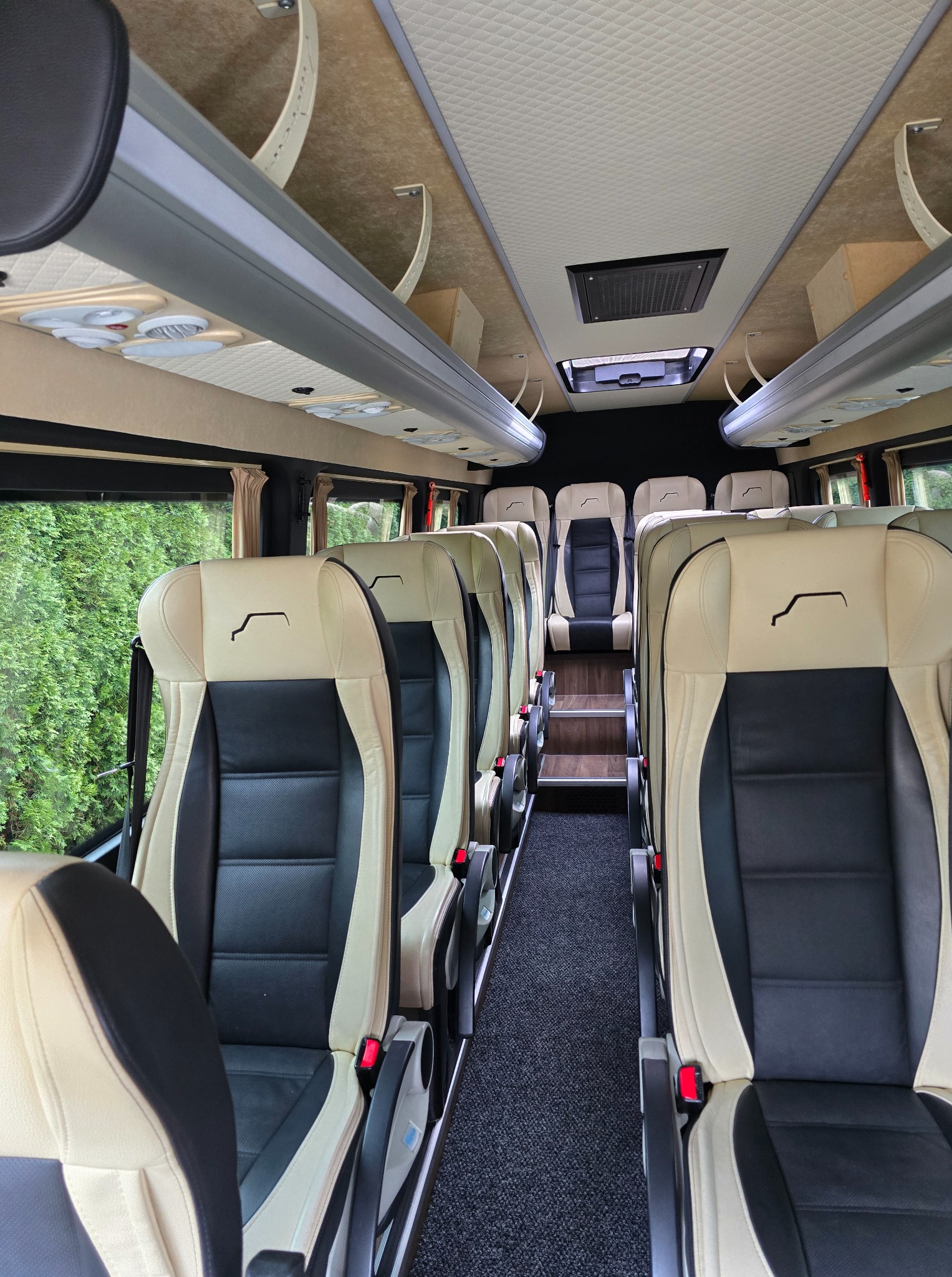 Wnętrze luksusowego busa z beżowo-czarnymi skórzanymi fotelami, oświetleniem sufitowym i widokiem zieleni za oknem, sugerujące komfortowy przewóz osób.