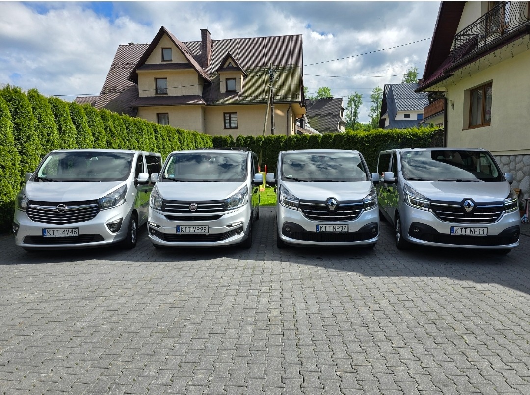 Flota srebrnych vanów (Opel, Fiat, Renault) zaparkowana rzędem na brukowanym podjeździe przed budynkiem i żywopłotem. Tablice rejestracyjne KTT.