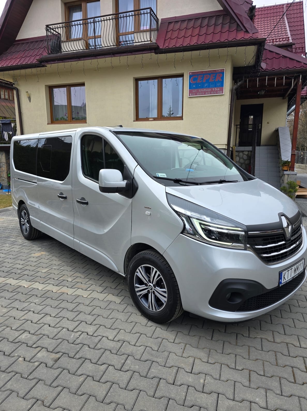 Srebrny Renault Trafic z ciemnymi szybami parkuje przed budynkiem z szyldem 'CEPER'. Widoczny numer rejestracyjny KTT WM11. Usługa przewozu osób.