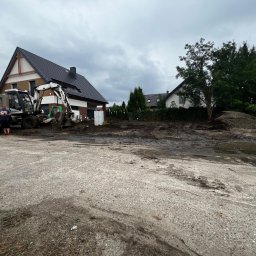 Bracia ogrodnicy. - Przygotowanie terenu pod budowę: koparka, wywrotka i operator na placu budowy. Widoczne fundamenty domu i sąsiednia zabudowa w tle, pochmurne niebo.