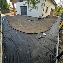 Bracia ogrodnicy. - Przygotowanie terenu pod ścieżkę wokół domu: czarna agrowłóknina, obrzeża i wyrównana ziemia. W tle jasny dom z elementami drewna i pracująca osoba.