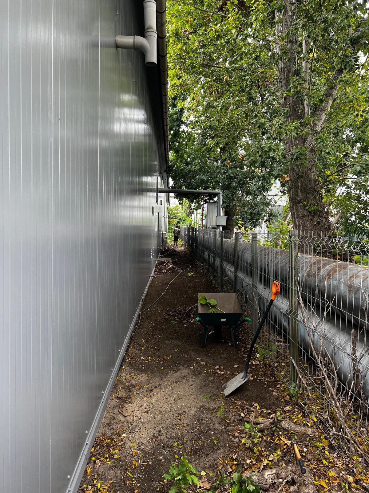 Wąskie przejście wzdłuż budynku, z wózkiem ogrodowym i łopatą. Widoczne rury i siatka ogrodzeniowa. Osoba w oddali, sugerująca prace porządkowe na zaniedbanym terenie.