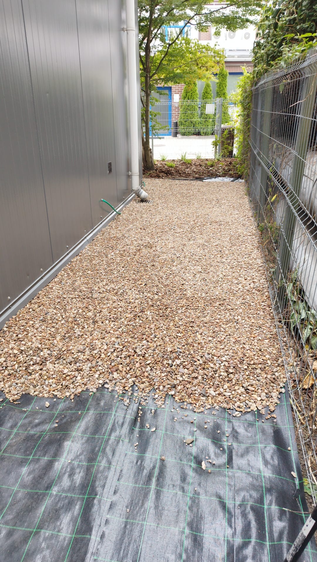 Wąskie przejście wyłożone drobnymi kamieniami na czarnej geowłókninie z zieloną siatką, obok metalowego garażu i ogrodzenia z siatki.