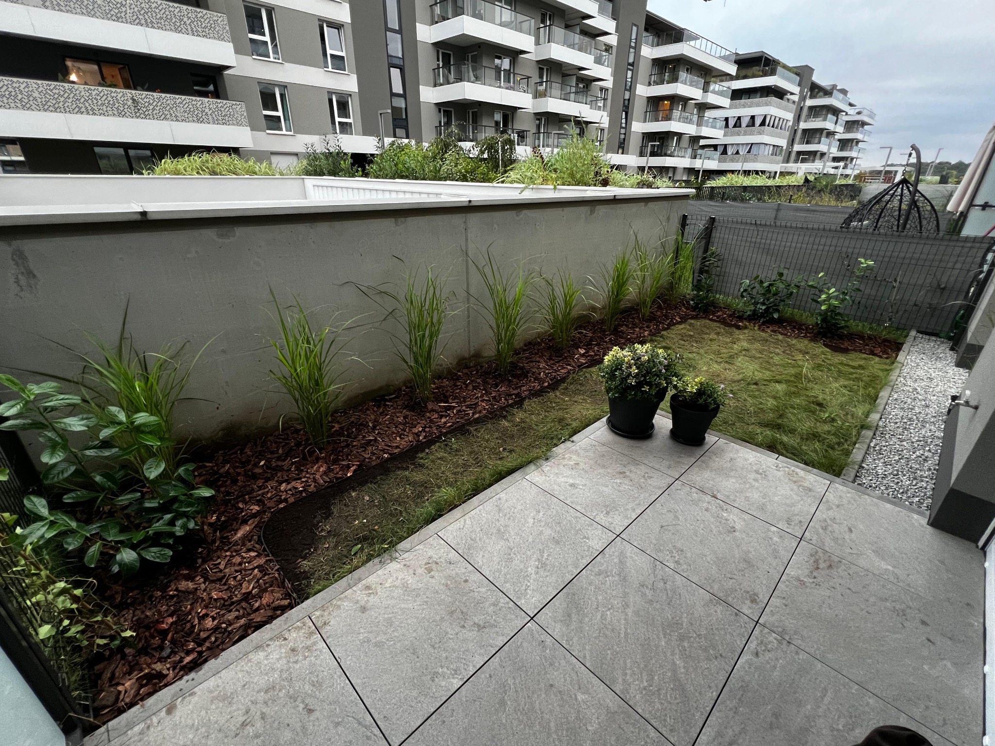 Nowoczesny ogród na balkonie z trawami ozdobnymi, korą i dwoma donicami z kwiatami na tle apartamentowca. Widok z góry na szare płytki balkonowe.