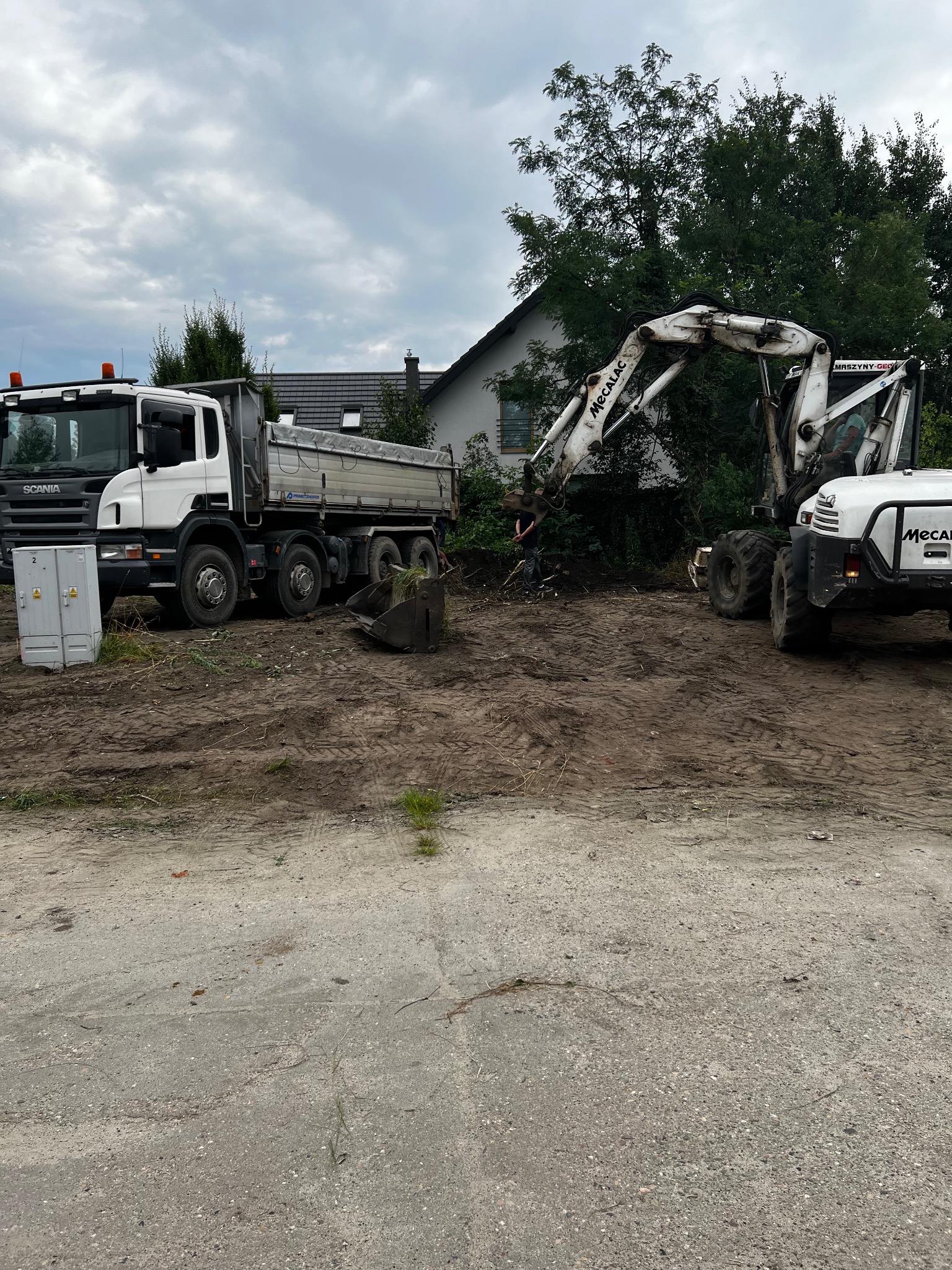 Ciężarówka i koparka Mecalac na placu budowy, przygotowującym teren pod inwestycję. W tle dom jednorodzinny i pochmurne niebo.