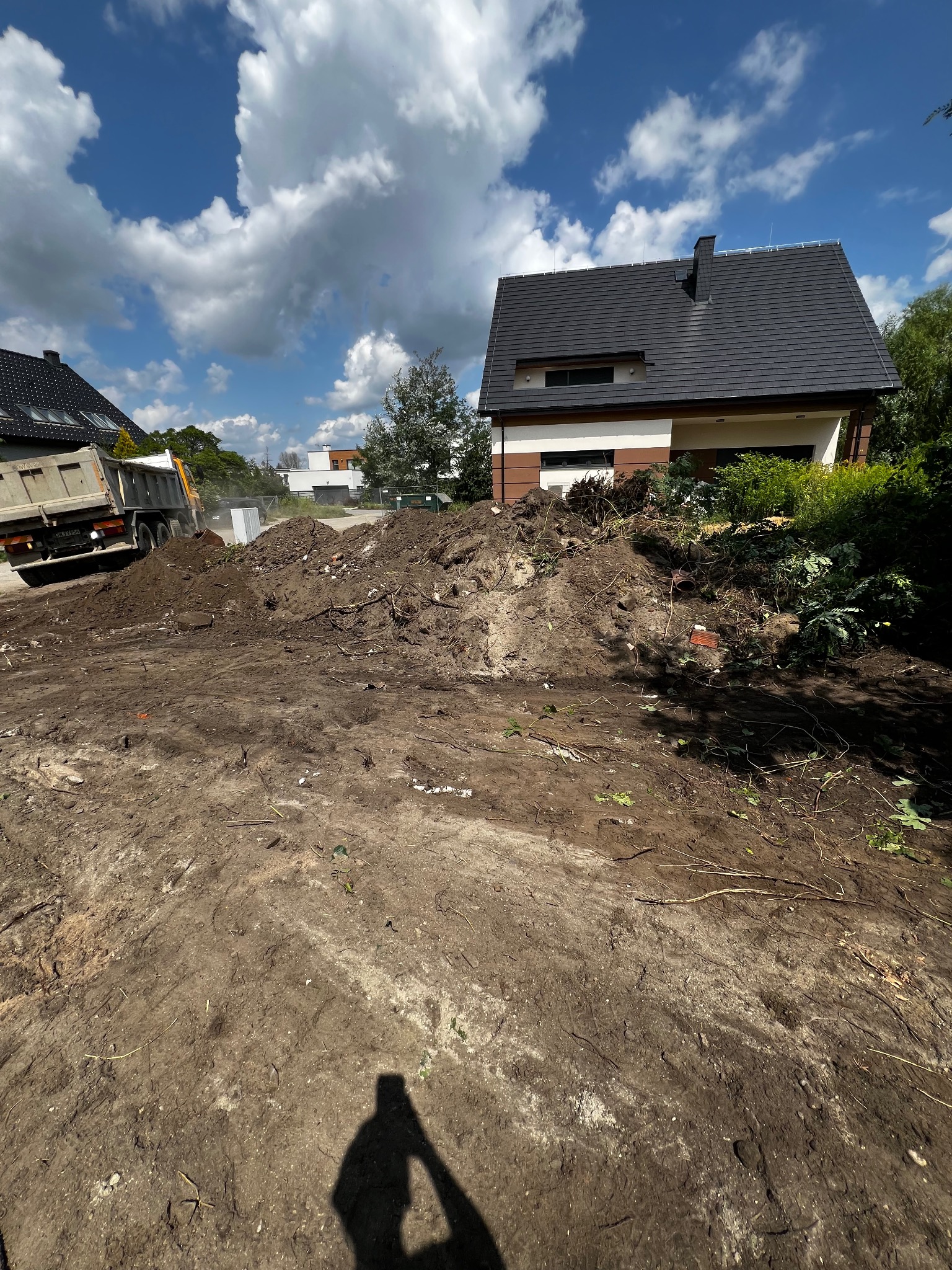 Wyładunek ziemi i gruzu z ciężarówki na działce przed domem z brązowo-białą elewacją. W tle błękitne niebo z obłokami. Cień fotografa w dolnej części kadru.