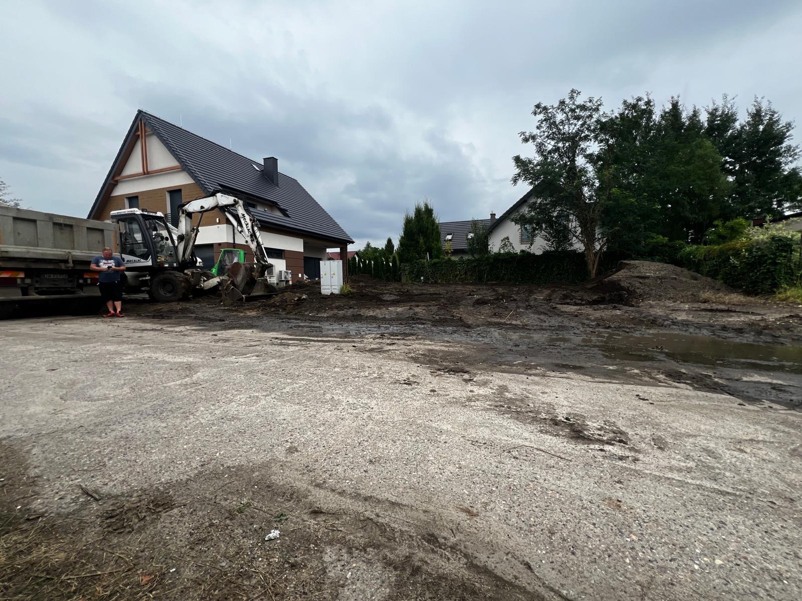 Przygotowanie terenu pod budowę: koparka, wywrotka i operator na placu budowy. Widoczne fundamenty domu i sąsiednia zabudowa w tle, pochmurne niebo.