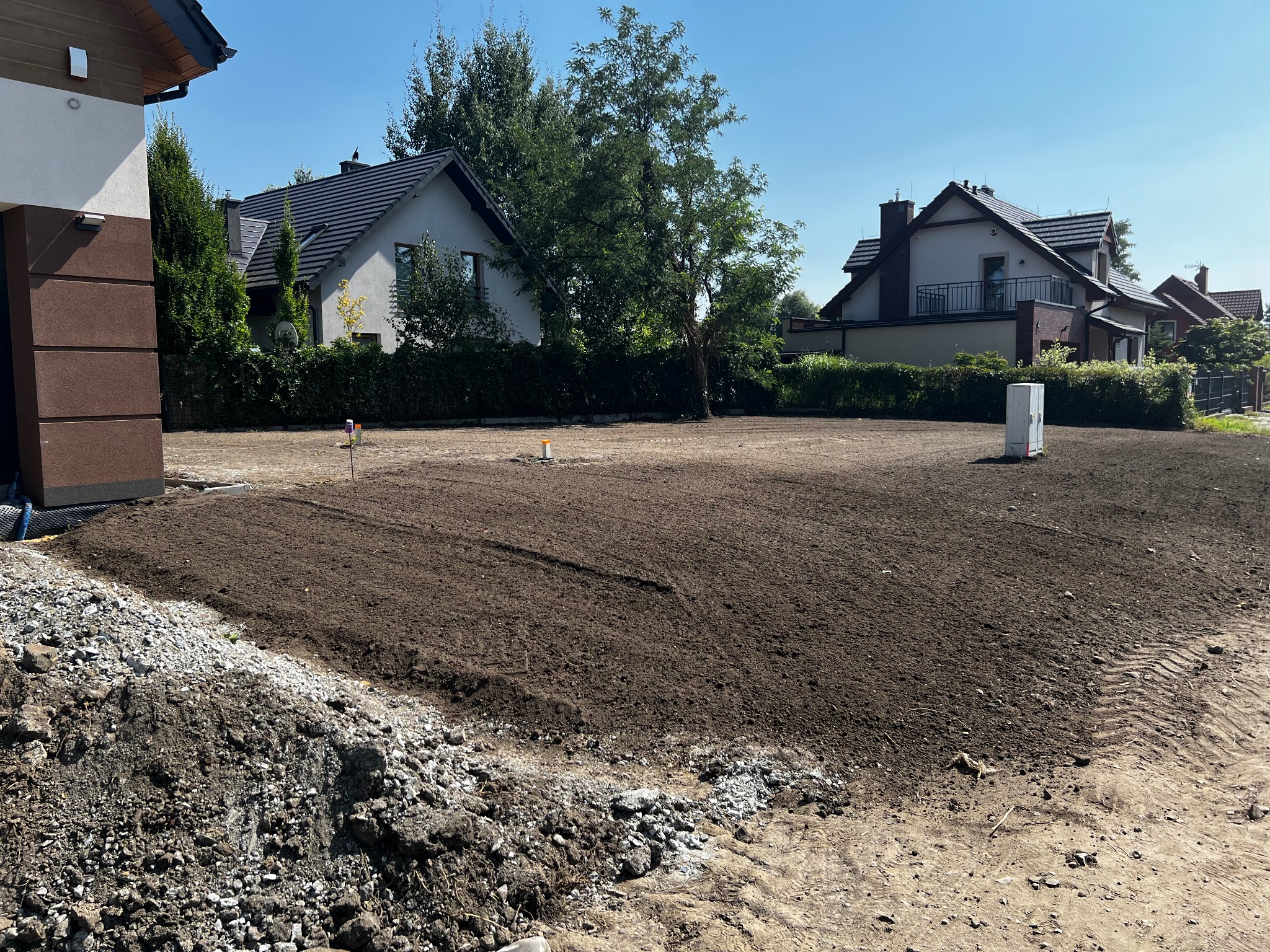 Przygotowany teren pod przyszły ogród z widocznymi domami w tle. Świeżo przekopana ziemia czeka na dalsze prace. Widoczne ślady opon i gruz.