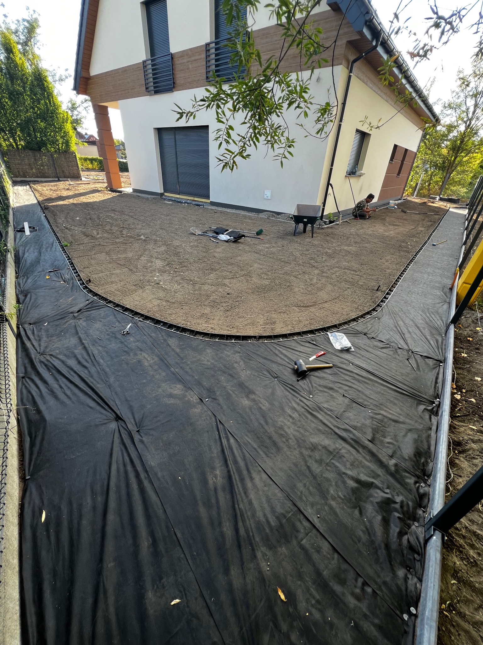 Przygotowanie terenu pod ścieżkę wokół domu: czarna agrowłóknina, obrzeża i wyrównana ziemia. W tle jasny dom z elementami drewna i pracująca osoba.