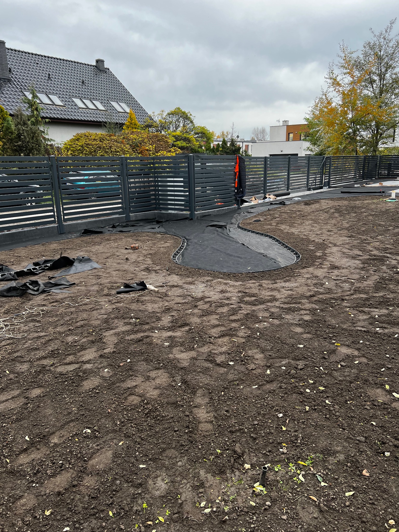 Przygotowanie terenu pod trawnik z rolki we Wrocławiu. Widoczne czarne maty i obrzeża rabat na przygotowanej ziemi. W tle nowoczesne ogrodzenie i dom.