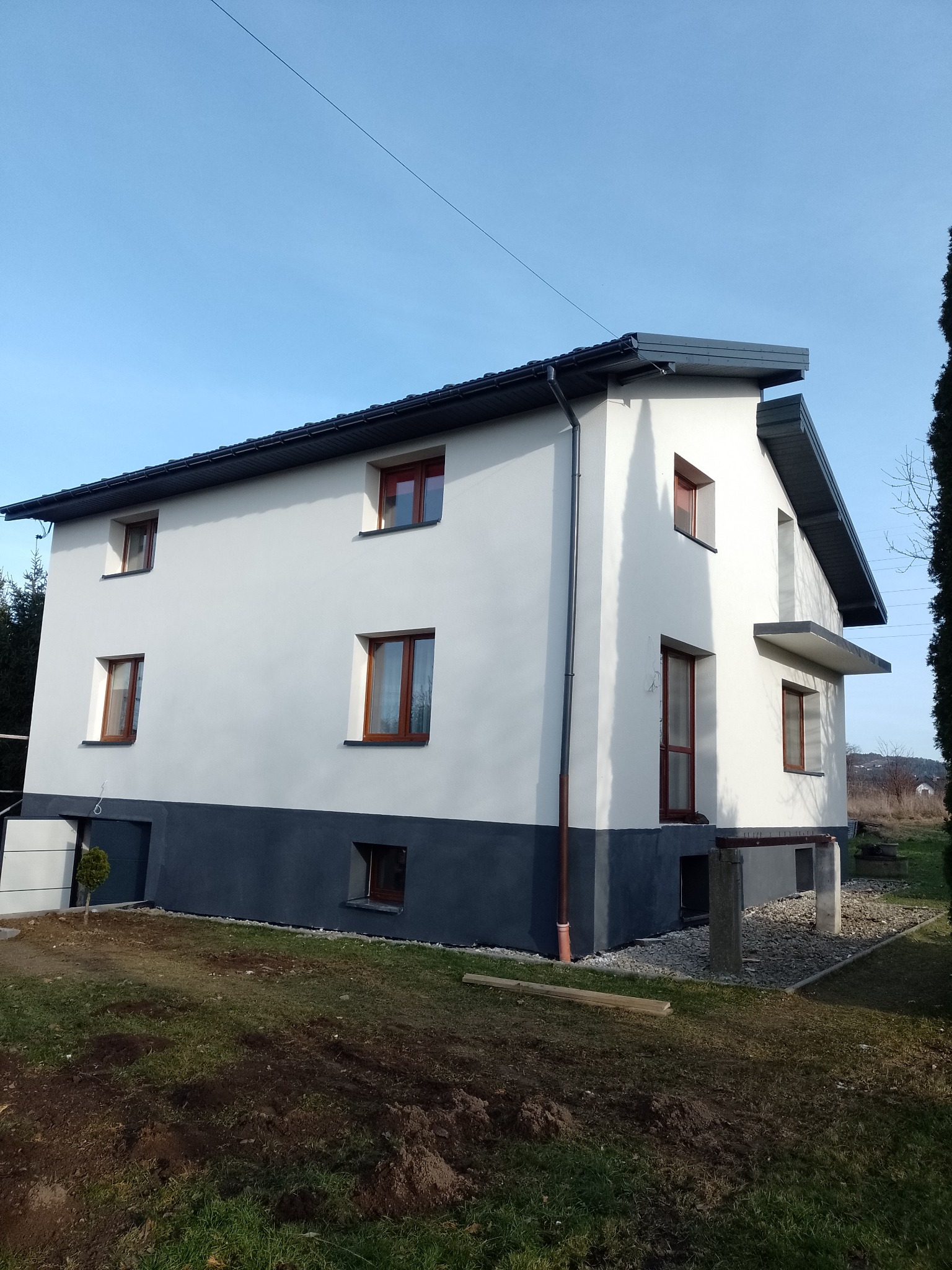 Dwukondygnacyjny dom po renowacji elewacji w bieli i szarości, z brązowymi oknami. Widok z boku na tle błękitnego nieba i zieleni ogrodu. Nowoczesne wykończenie.