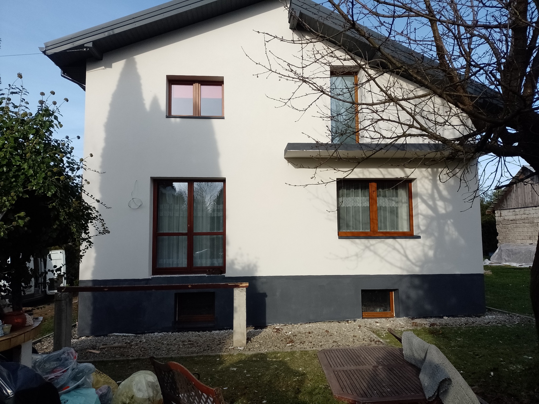 Dwukolorowa elewacja domu jednorodzinnego z drewnianą stolarką okienną. Biała ściana kontrastuje z szarym cokołem. Widoczne drzewo bez liści i fragment wyposażenia ogrodu.