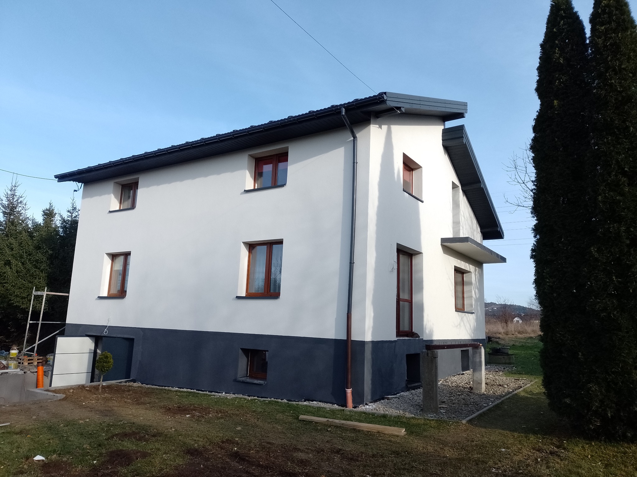 Dwukondygnacyjny dom z białą elewacją i ciemnoszarą podmurówką w słoneczny dzień. Widoczne brązowe okna, szara dachówka i elementy rusztowania w tle.