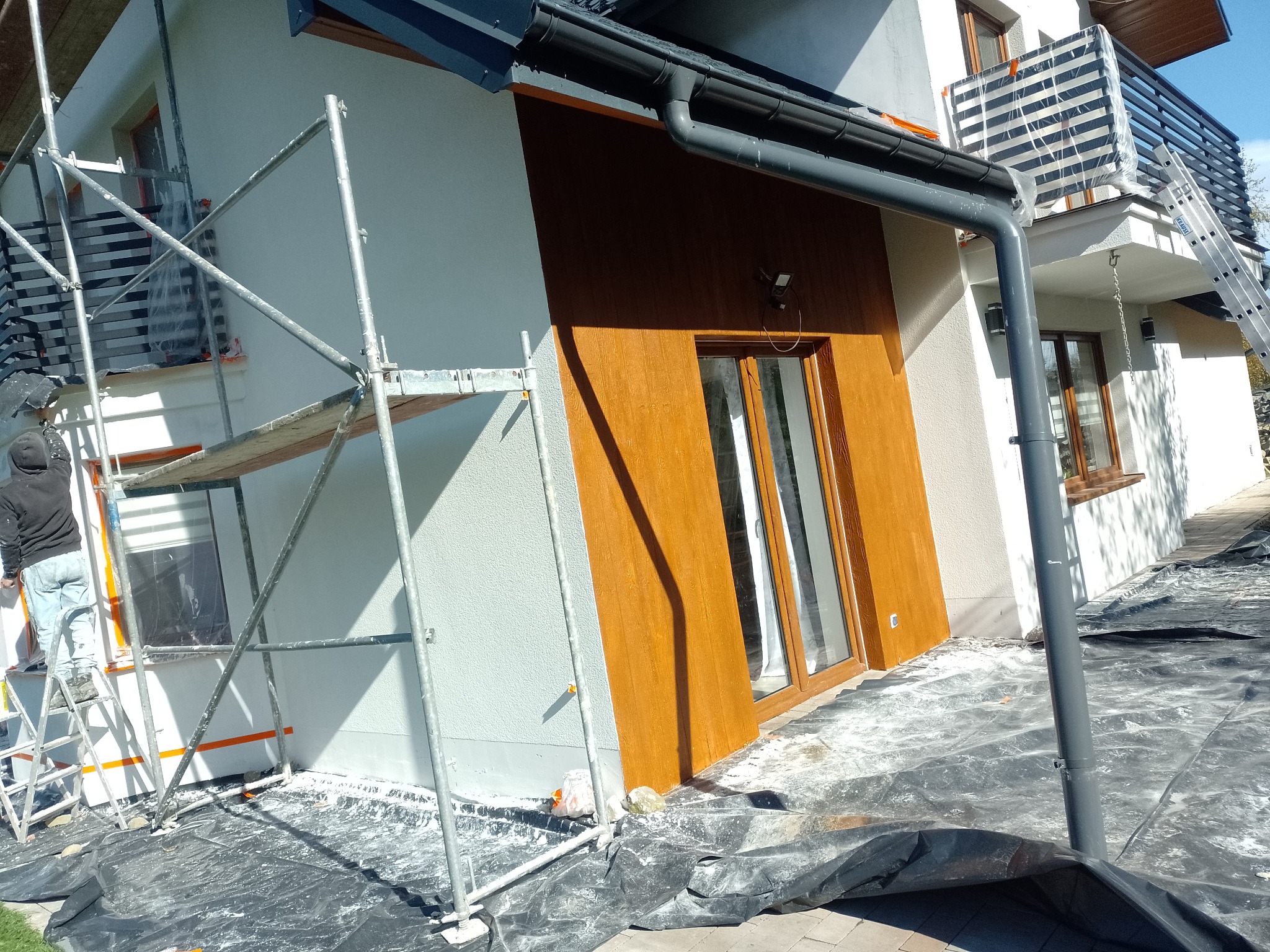 Prace elewacyjne w toku: ocieplony budynek z drewnianą okładziną, rusztowania i pracownik na drabinie. Ochronna folia na kostce brukowej.