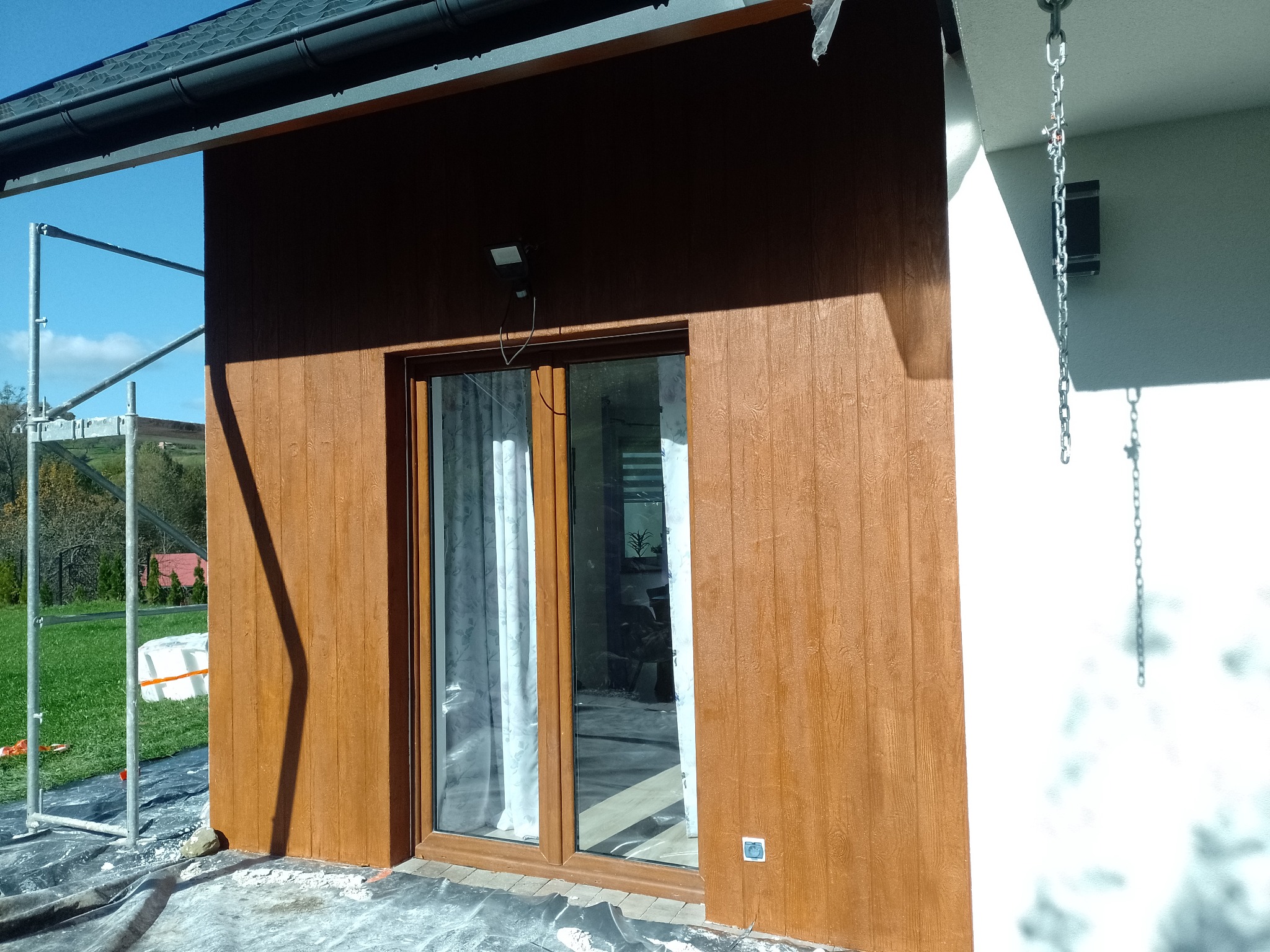 Wykończona elewacja domu z drewnopodobną okładziną, widoczne rusztowanie i teren przygotowany do dalszych prac wykończeniowych.