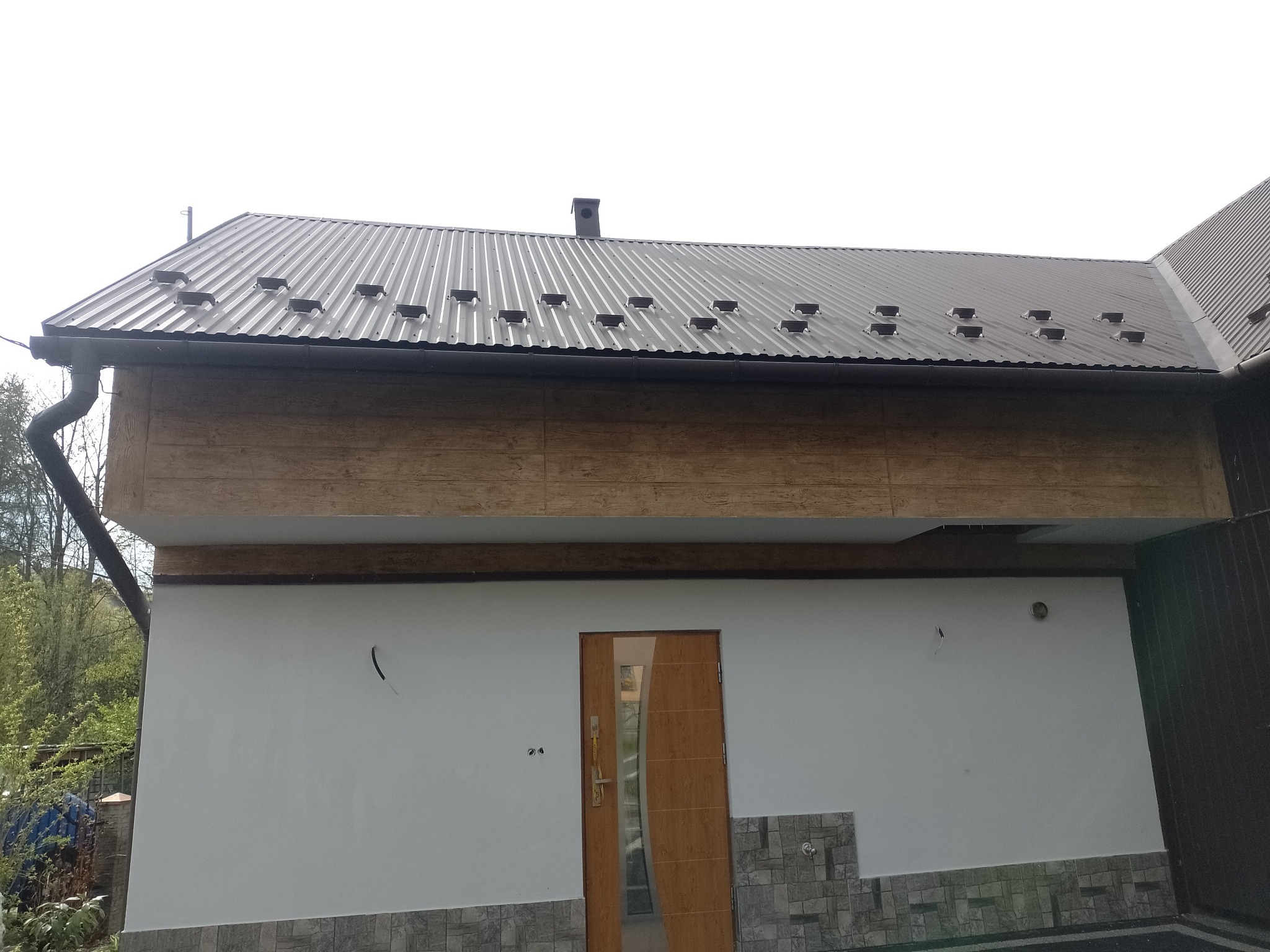 Wykończenie elewacji domu w Łącku: biała ściana z drewnianą wstawką i brązowym dachem z blachy trapezowej. Widoczny fragment rynny i drzwi wejściowe.