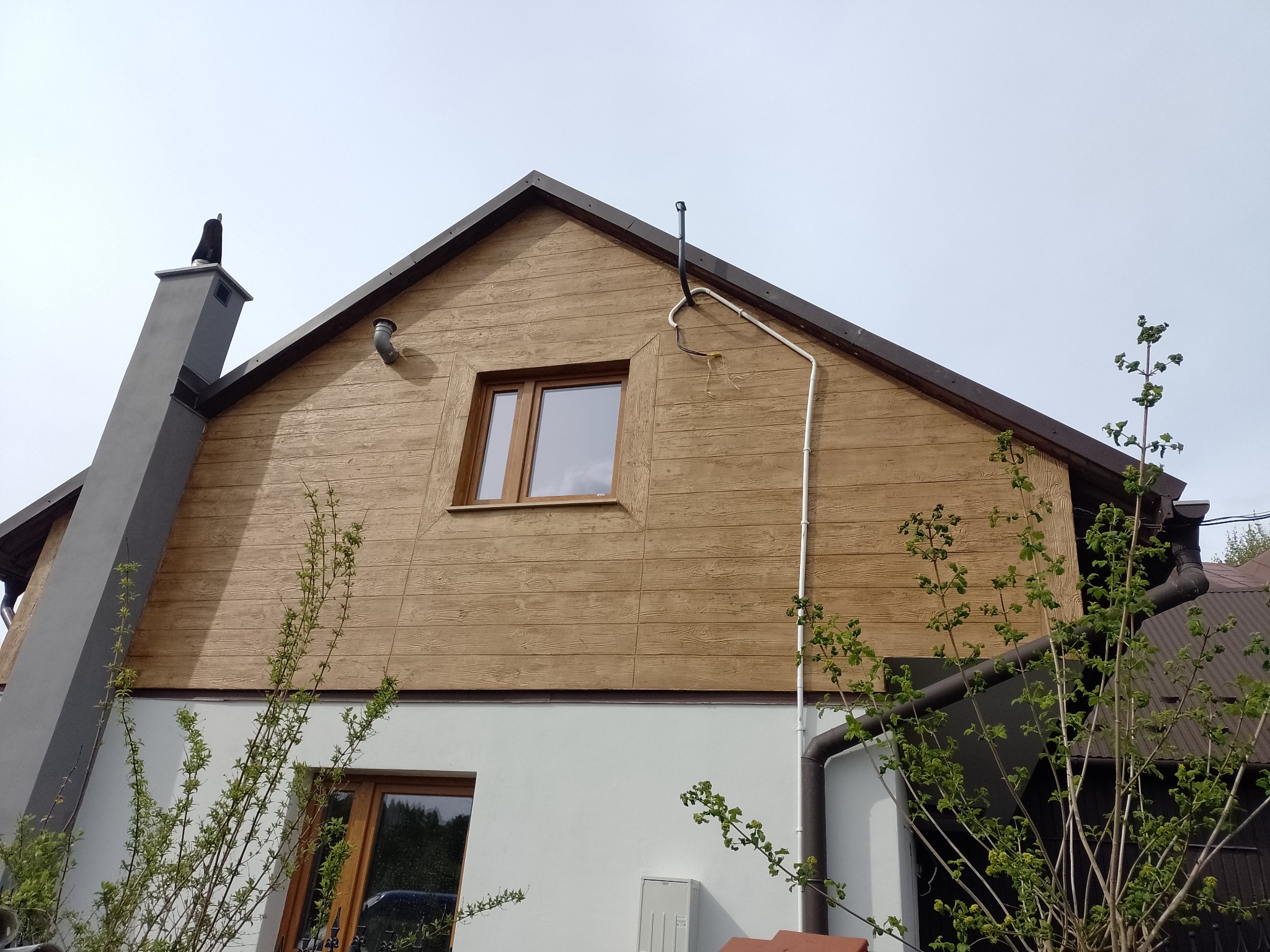 Dom z drewnianą elewacją i szarym kominem, widoczny z dołu pod kątem. Elewacja imituje drewno, okna z brązowymi ramami. Widoczne rynny i fragmenty roślin.