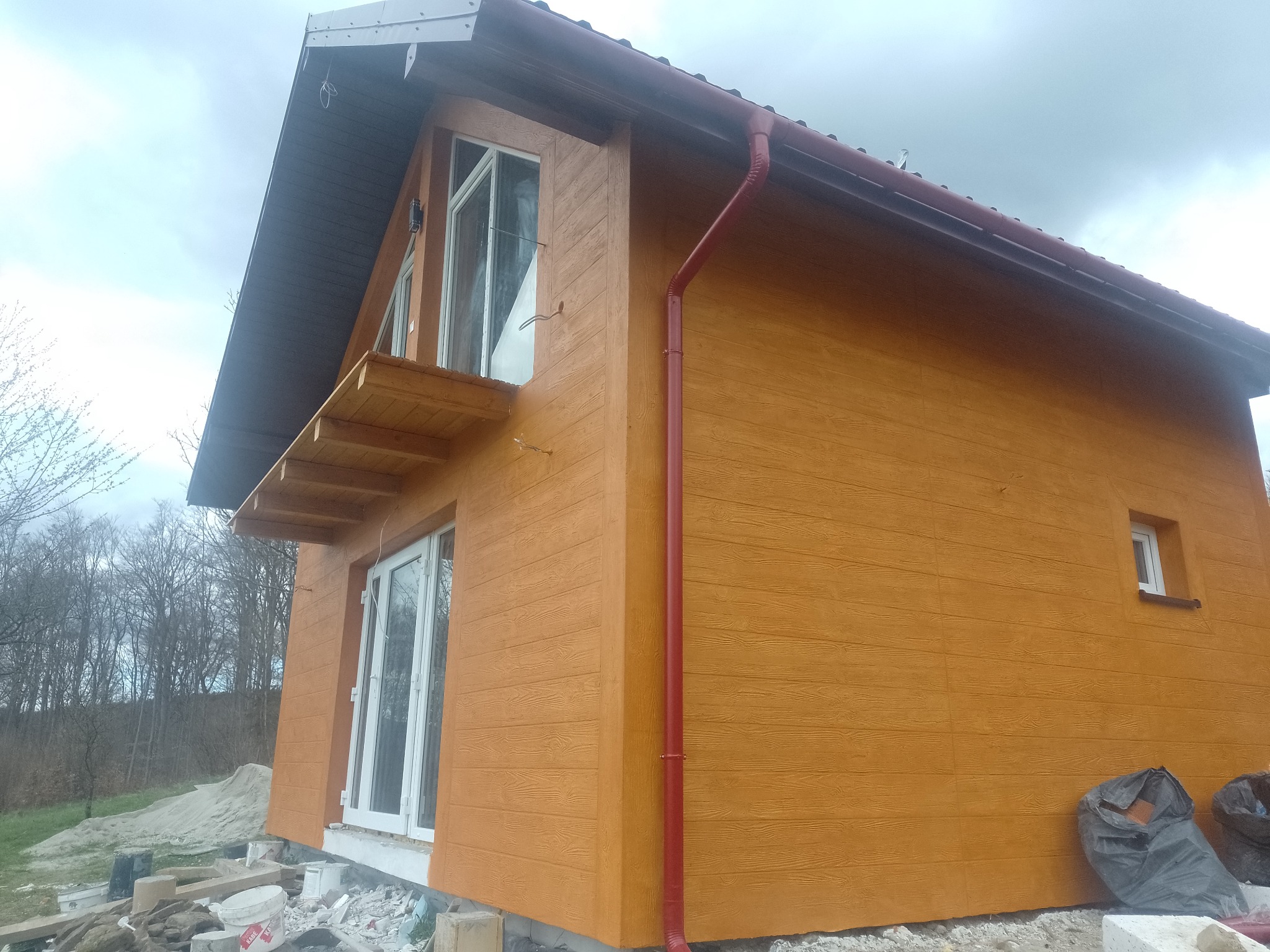 Wykończona elewacja domu jednorodzinnego w odcieniach brązu, z widocznymi oknami i elementami dachu. Na pierwszym planie materiały budowlane.