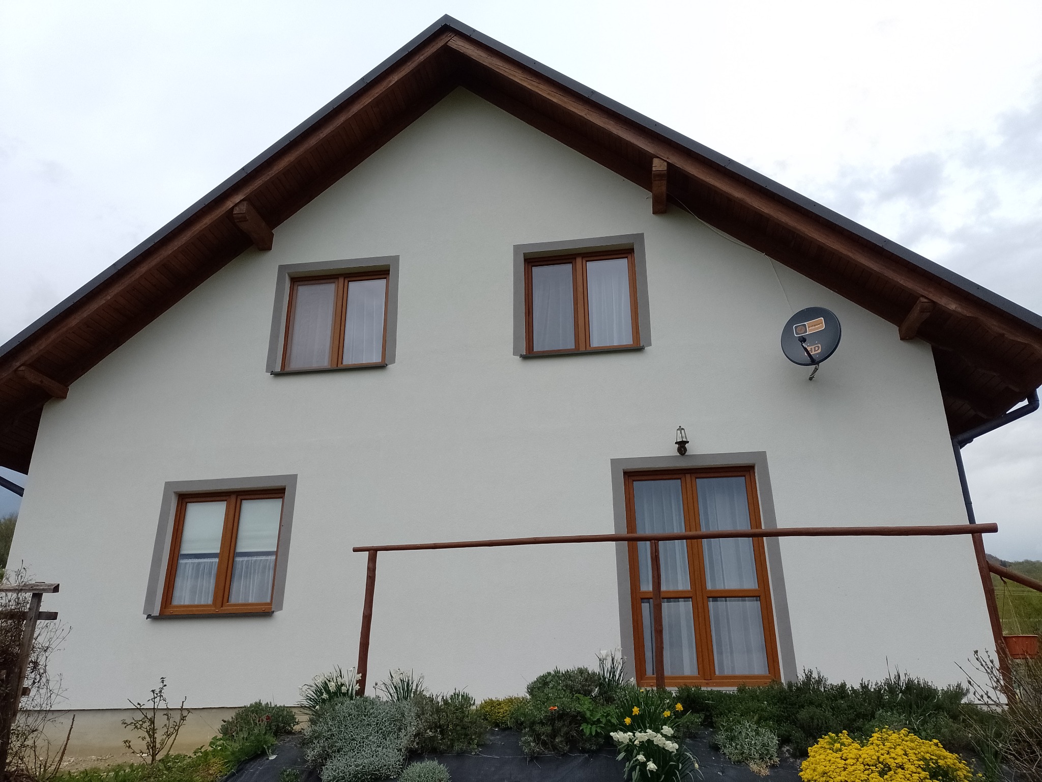 Biała elewacja domu jednorodzinnego z drewnianymi oknami i brązowym dachem. Widoczny fragment ogrodu z niską barierką. Antena satelitarna na ścianie.