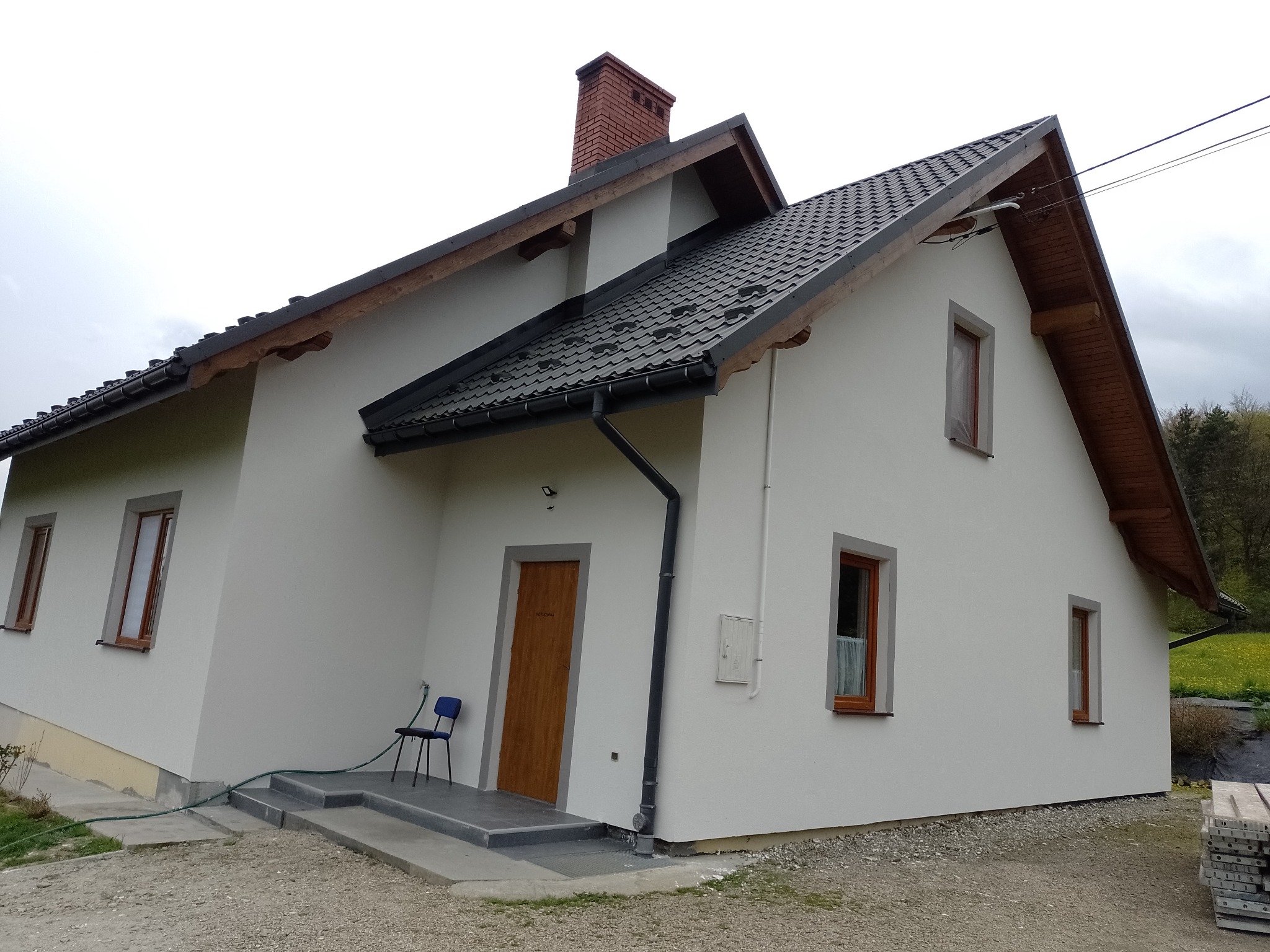 Biały dom jednorodzinny z ciemną dachówką i drewnianymi oknami, widok z boku, z krzesłem przy wejściu i stertą desek w tle.