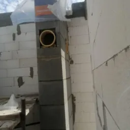 Wznoszenie komina z pustaków szarych, z widocznymi otworami na rury wentylacyjne. Na górze folia zabezpieczająca. Białe bloczki ściany w tle. Budowa w toku.