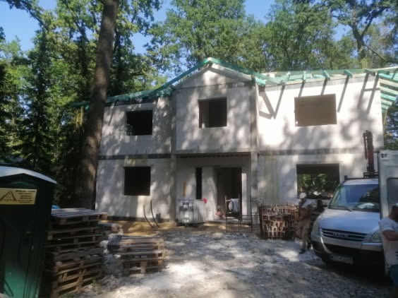 Dwukondygnacyjny dom w stanie surowym otwartym, z widoczną konstrukcją dachu z drewna. W tle drzewa, na pierwszym planie palety i pojazd budowlany.