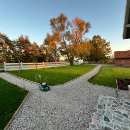 Green Tech - Widok na świeżo założony trawnik z białym ogrodzeniem i ścieżką z kamyczków, gdzie stoi zagęszczarka wibracyjna. W tle drzewa i budynek z czerwonej cegły.