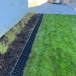 Świeżo założony trawnik z systemem drenażowym i rabatą z ozdobnymi trawami, widok z góry. Na drugim planie fragment domu z klimatyzacją.