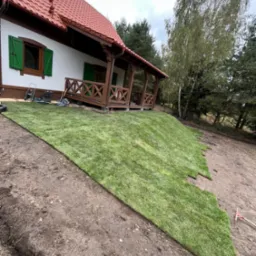 Układanie trawy z rolki na zboczu przy domu z drewnianą werandą. Widoczne narzędzia ogrodnicze i częściowo ułożona trawa. Tło stanowią drzewa.