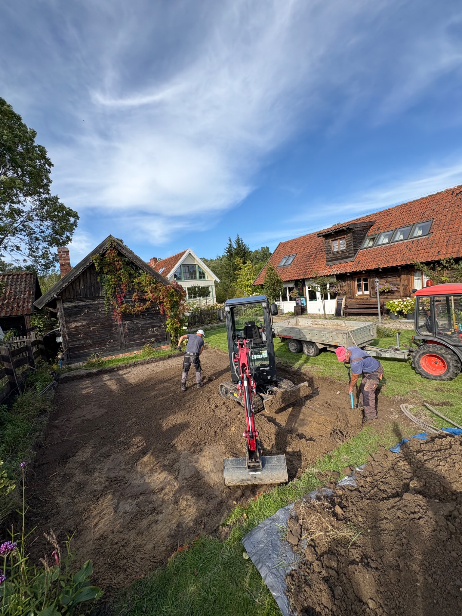 Prace ziemne w ogrodzie: mini-koparka, traktor z przyczepą i dwóch pracowników wyrównujących teren pod budowę, w tle zabudowa wiejska.