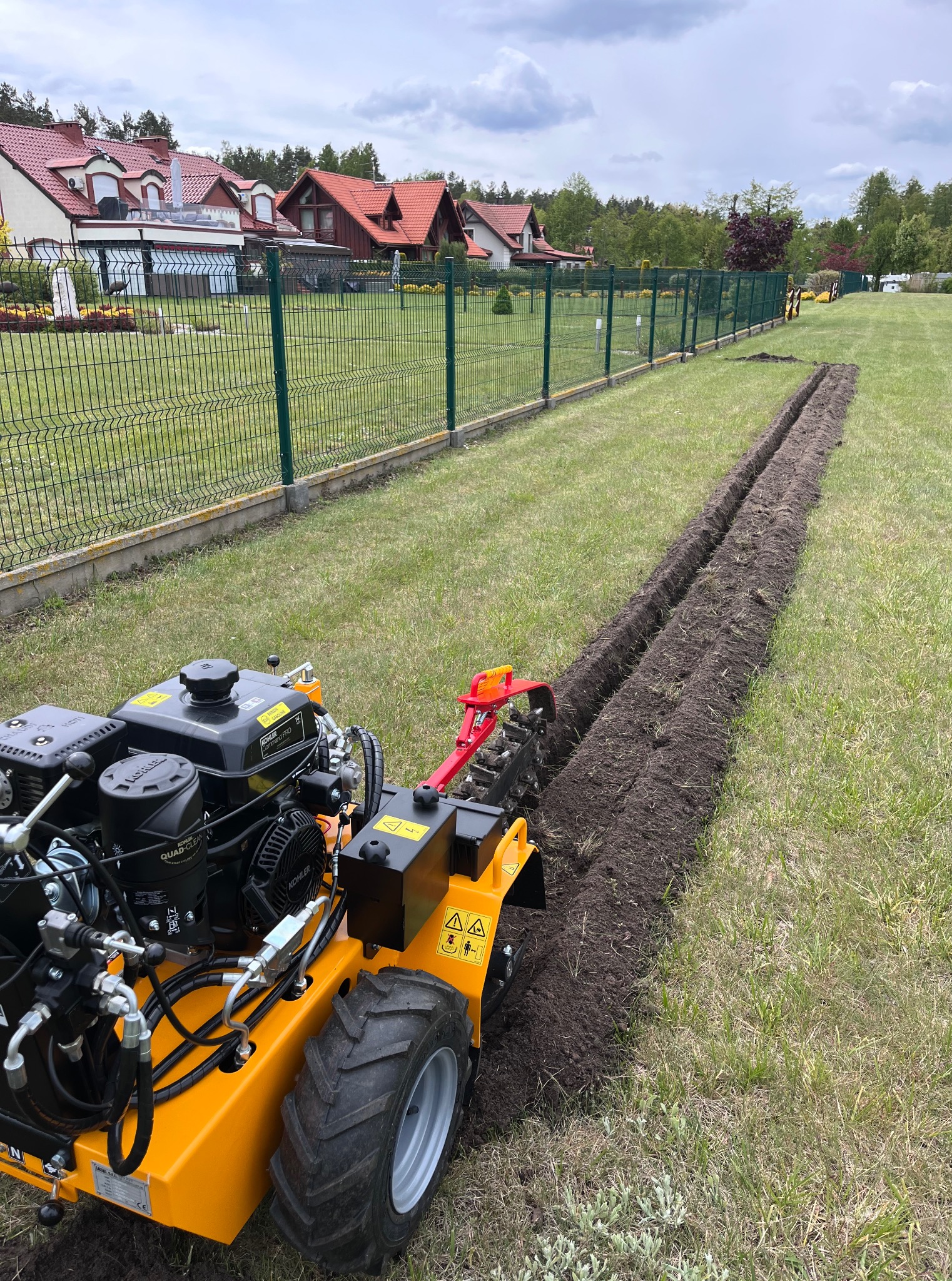 Żółta glebogryzarka wykopuje wąski rów w trawniku wzdłuż ogrodzenia z siatki na tle zabudowań mieszkalnych pod pochmurnym niebem.