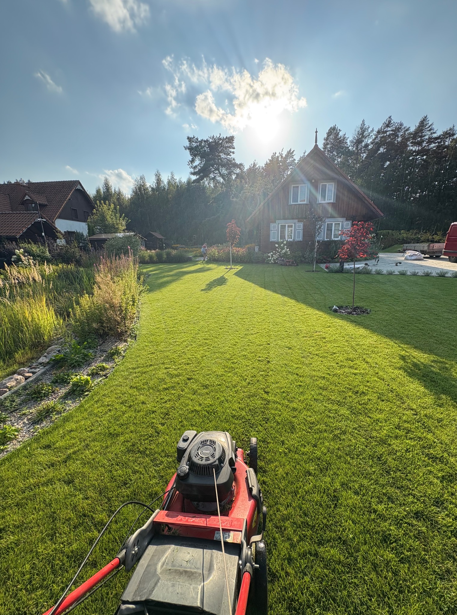 Czerwona kosiarka na tle idealnie skoszonego, zielonego trawnika przed domem z drewnianą elewacją w słoneczny dzień. W tle zieleń drzew i krzewów.
