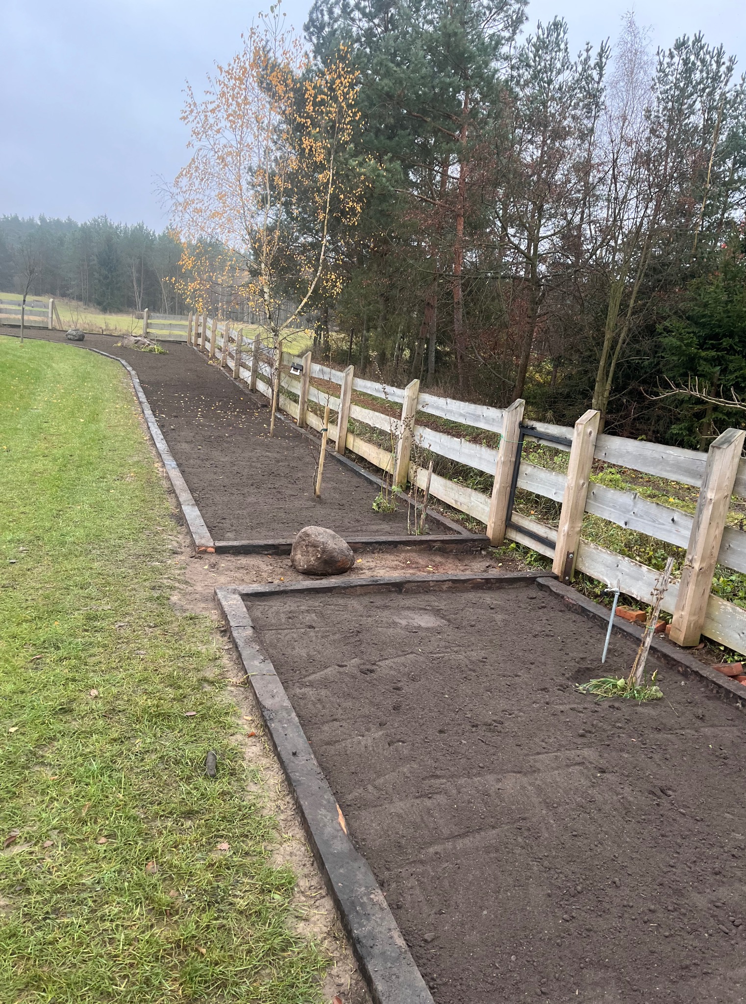 Przygotowane rabaty z obrzeżami i drewnianym płotkiem, gotowe do sadzenia w ogrodzie. Widoczne drzewka i duży kamień dekoracyjny. Pochmurny dzień.