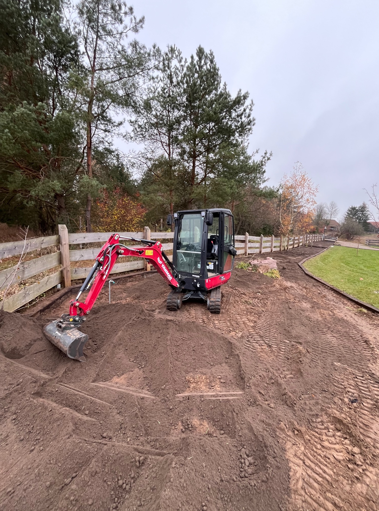 Czerwona mini koparka Yanmar w trakcie prac ziemnych przy drewnianym płocie na działce. Ślady opon na ziemi. Teren przygotowany pod dalsze prace.