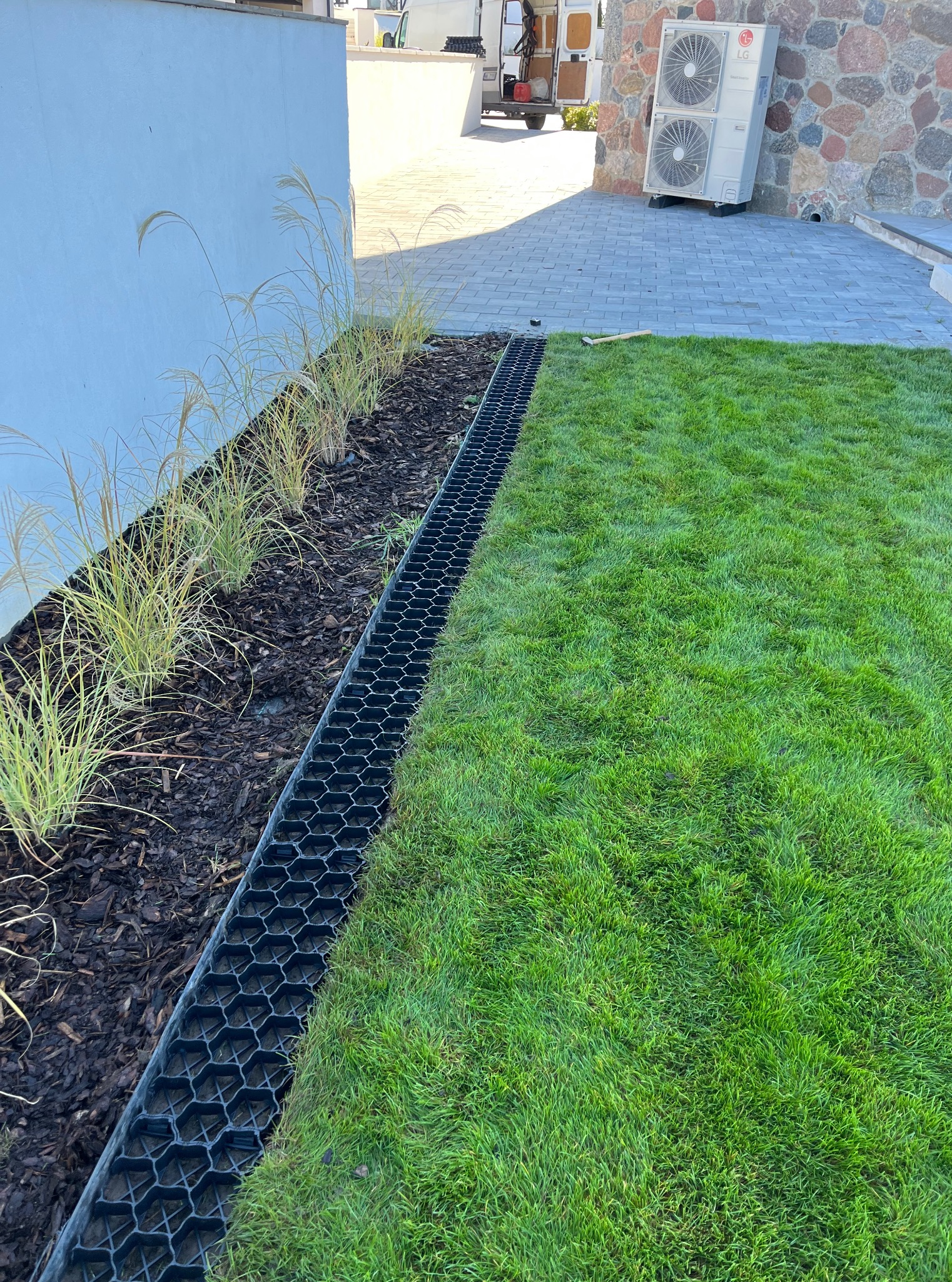 Świeżo założony trawnik z systemem drenażowym i rabatą z ozdobnymi trawami, widok z góry. Na drugim planie fragment domu z klimatyzacją.