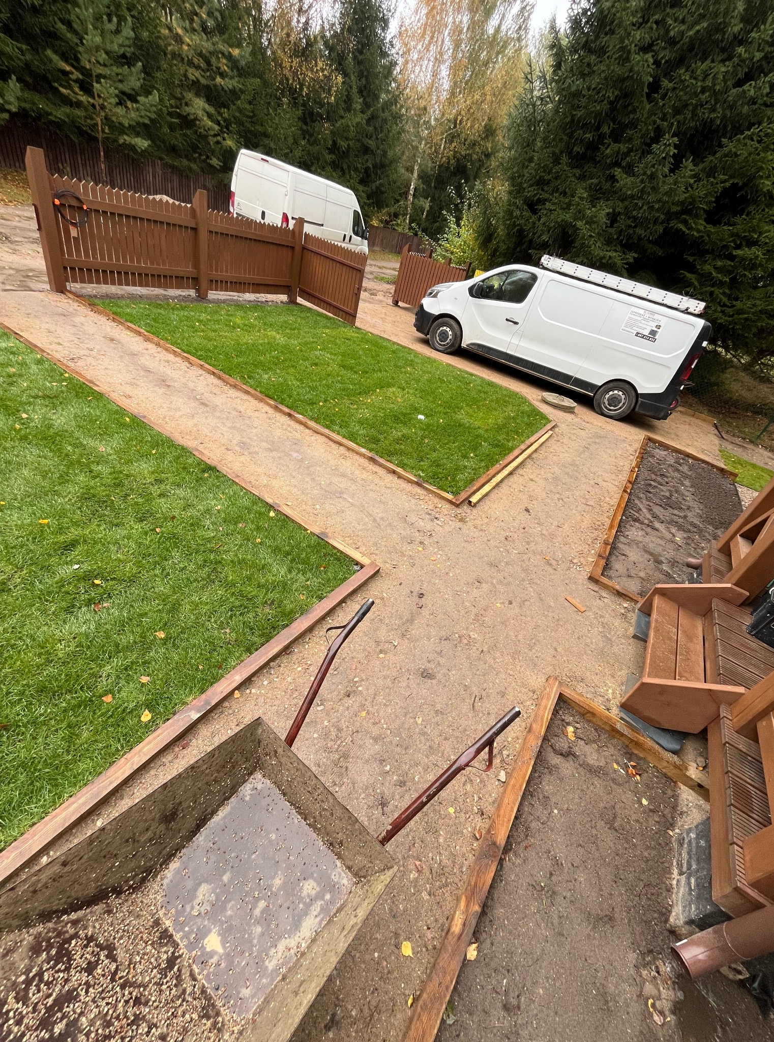 Fragment ogrodu w trakcie realizacji: ułożona trawa, obrzeża z drewna, taczka wypełniona materiałem i dwa busy w tle. Widok z góry.