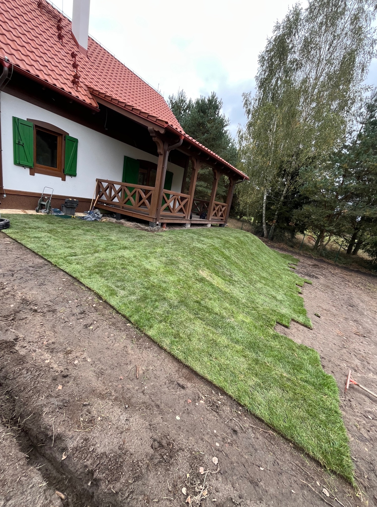 Układanie trawy z rolki na zboczu przy domu z drewnianą werandą. Widoczne narzędzia ogrodnicze i częściowo ułożona trawa. Tło stanowią drzewa.