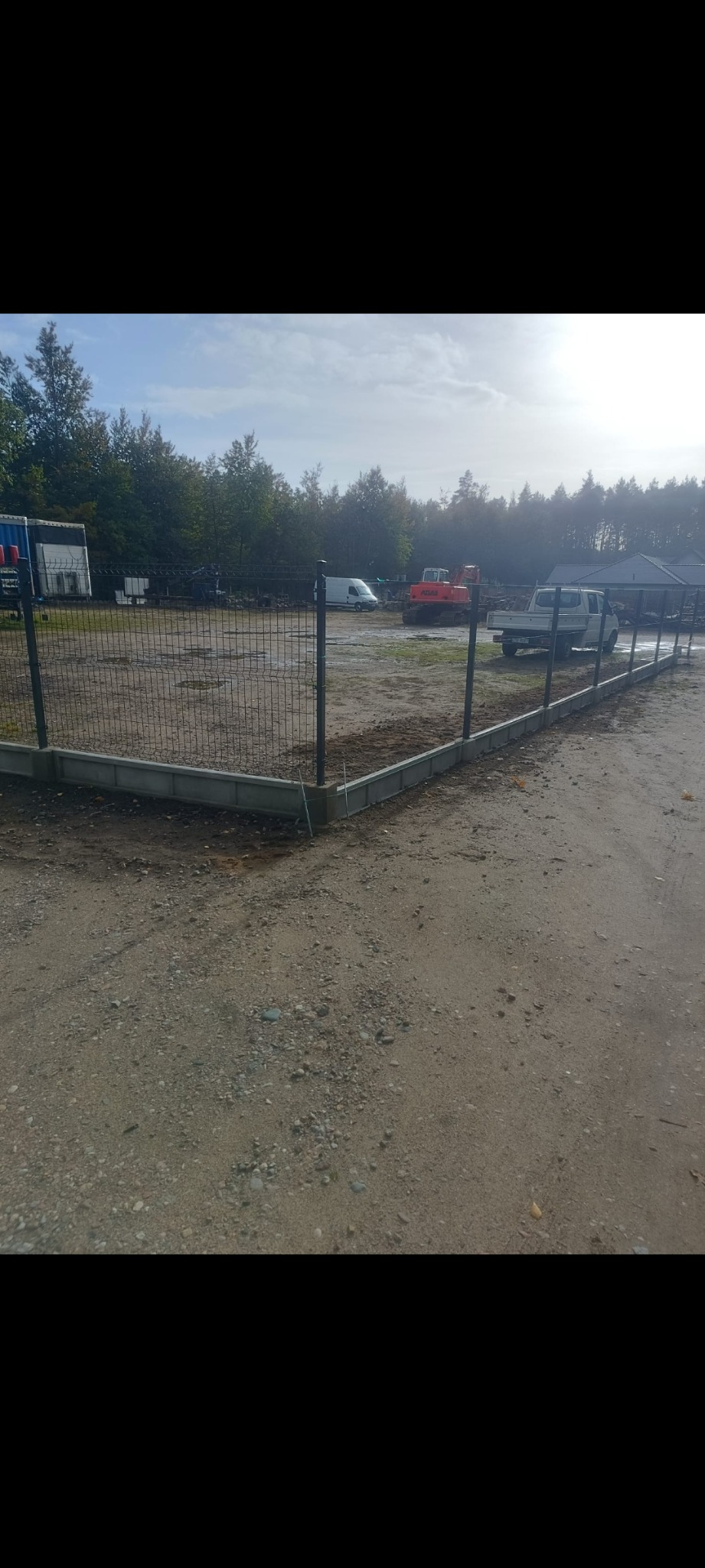 Nowoczesne ogrodzenie panelowe z podmurówką betonową, otaczające teren z widocznymi maszynami budowlanymi w tle. Ujęcie z poziomu gruntu, podkreślające solidność konstrukcji.