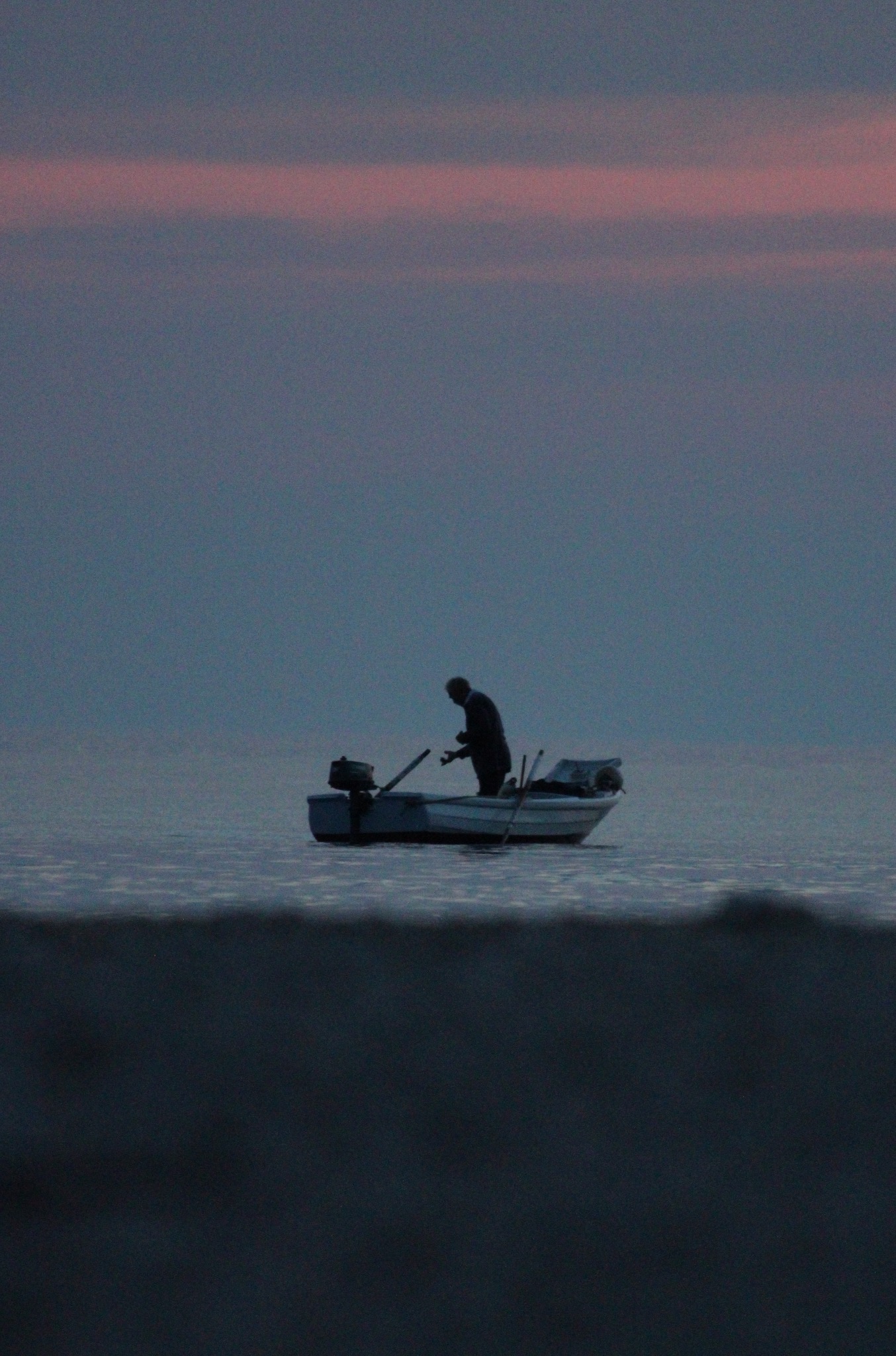 Silueta mężczyzny w łodzi na spokojnym jeziorze o zmierzchu, widok z niskiego kąta. W tle pas różowego nieba.