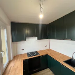 Nowoczesna kuchnia w odcieniach zieleni i drewna z widokiem z okna. Uwagę przykuwają ciemnozielone szafki z miedzianymi uchwytami i drewniany blat.