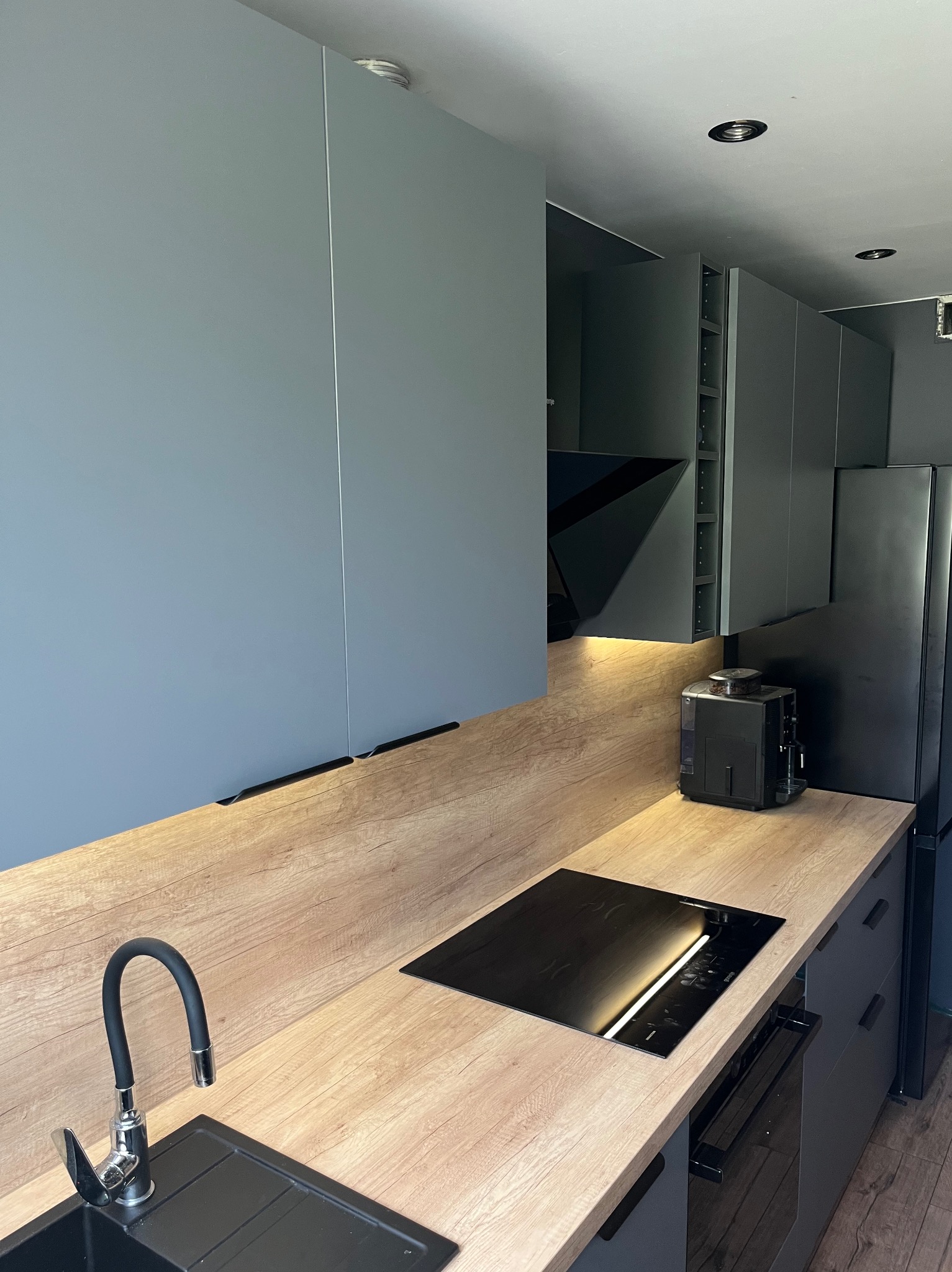 Nowoczesna kuchnia w szarościach z drewnianym blatem, czarną armaturą i sprzętami. Minimalistyczny design, podświetlane szafki górne, płyta indukcyjna.