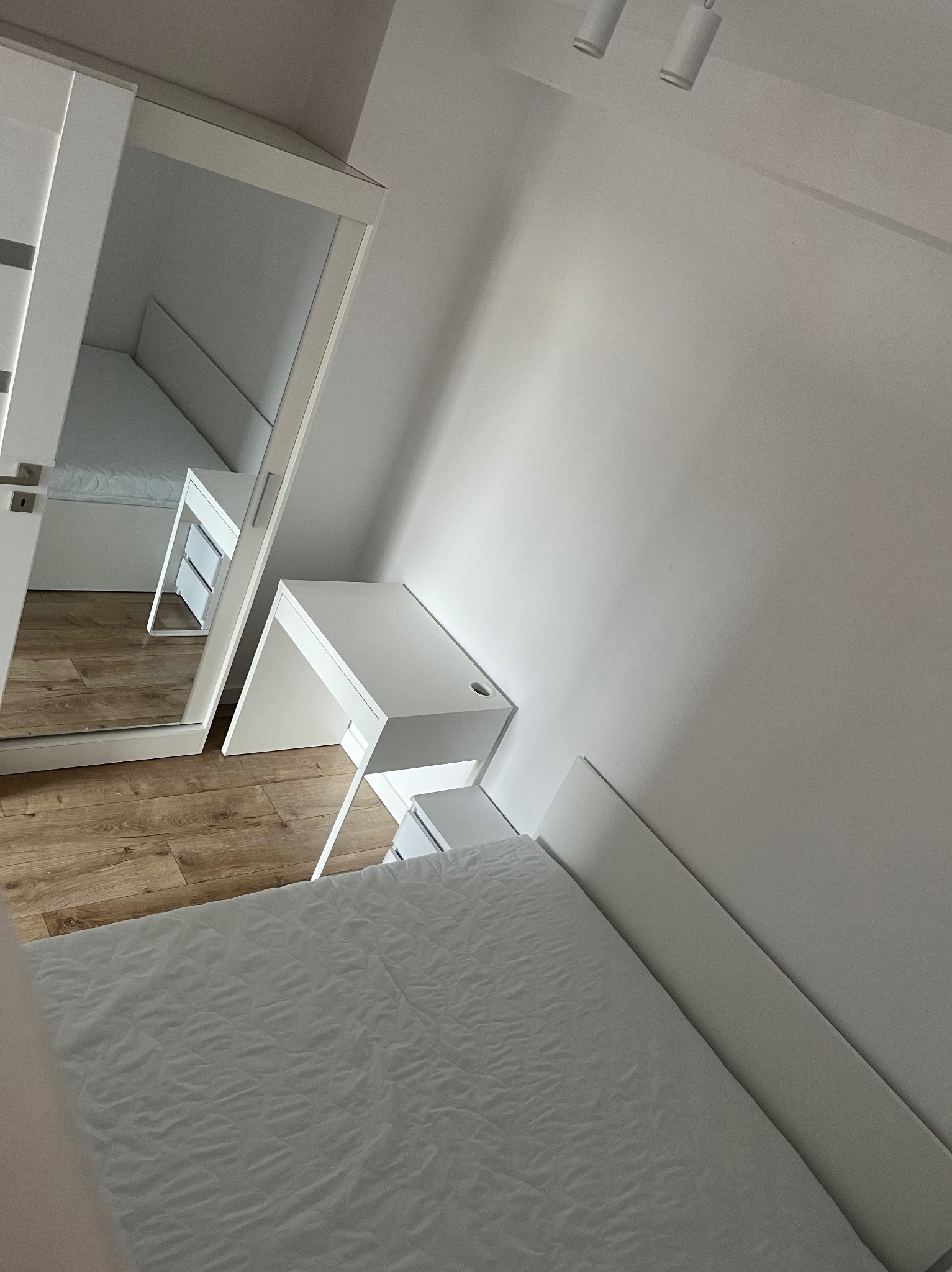 Minimalistyczna sypialnia w Piekary Śląskie z białymi meblami: łóżko, biurko i szafa z lustrem. Jasne drewno podłogi. Nowoczesne oświetlenie sufitowe.