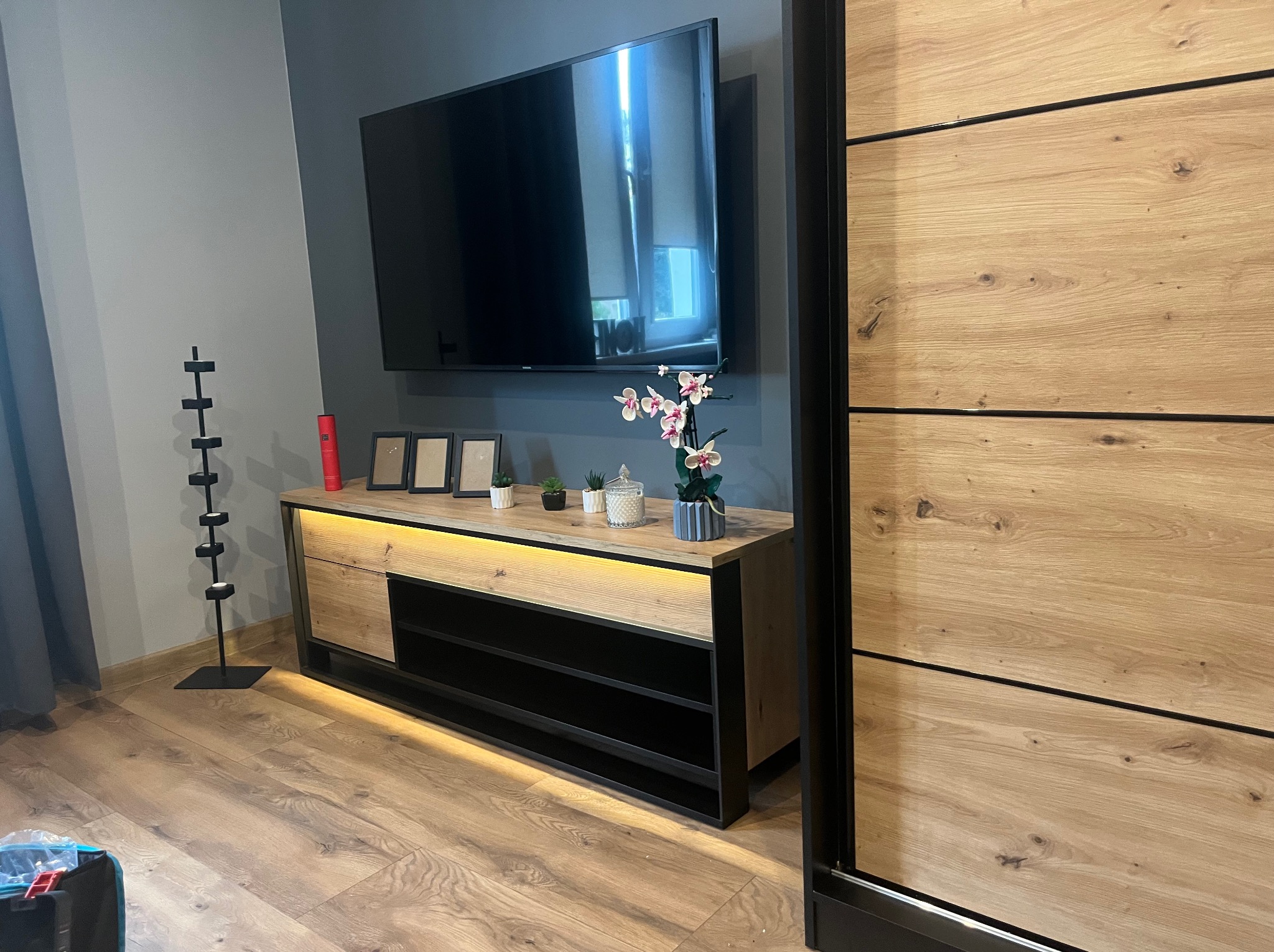 Nowoczesny salon z drewnianą komodą podświetloną LED, telewizorem na ścianie i dużą szafą przesuwną. Minimalistyczny design, ciepłe oświetlenie.