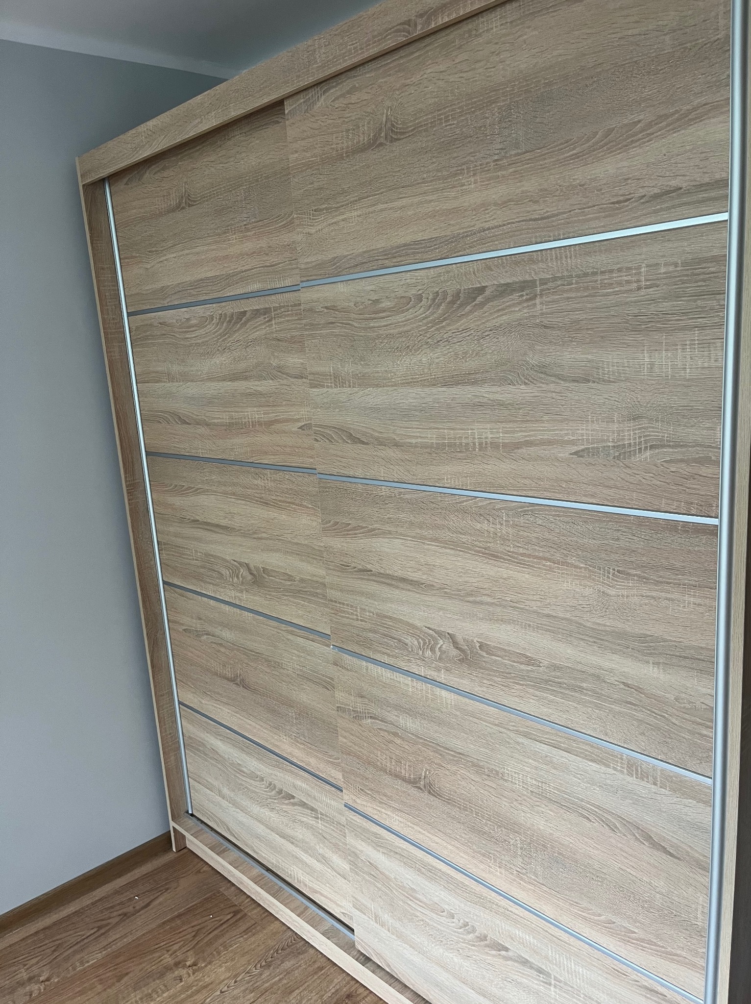 Duża, jasna szafa przesuwna z drewnianymi panelami i srebrnymi akcentami, stojąca na drewnianej podłodze przy szarej ścianie. Widoczne detale wykończenia.