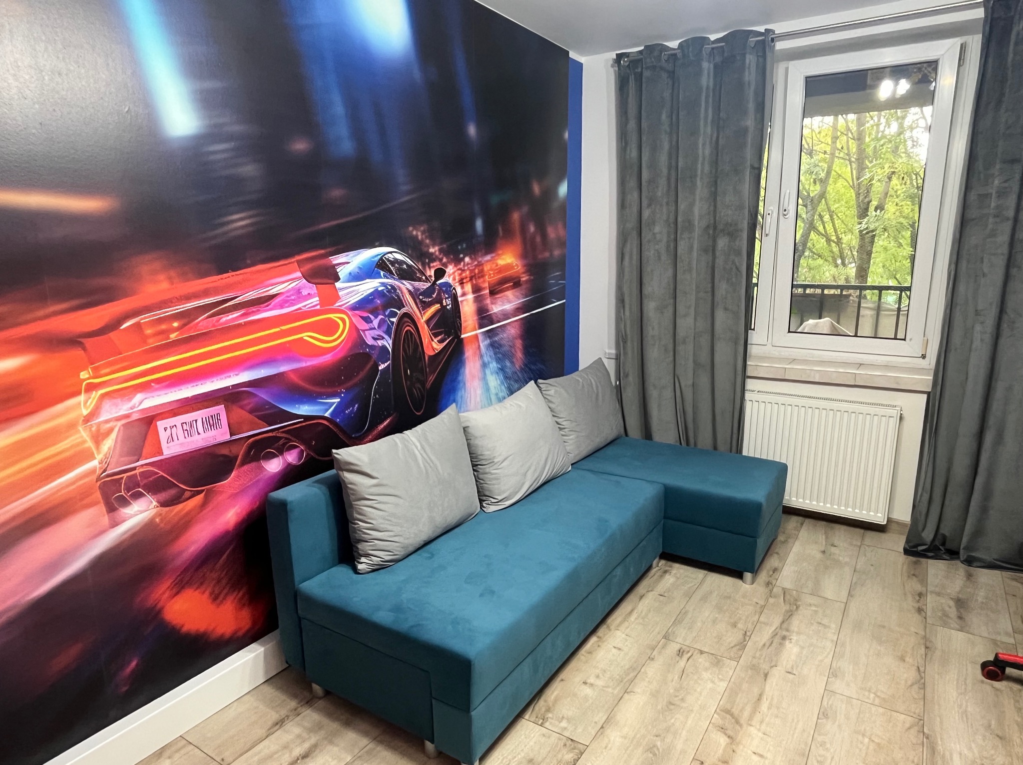 Pokój z turkusową sofą, szarymi zasłonami i fototapetą z motywem sportowego auta. Jasne panele podłogowe i okno z widokiem na zieleń.