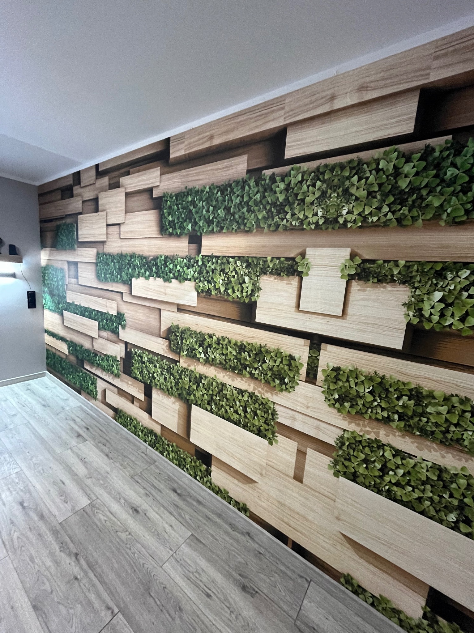 Nowoczesna ściana z drewnianymi panelami i zielonymi elementami roślinnymi, tworząca trójwymiarowy efekt. Jasna podłoga z paneli.