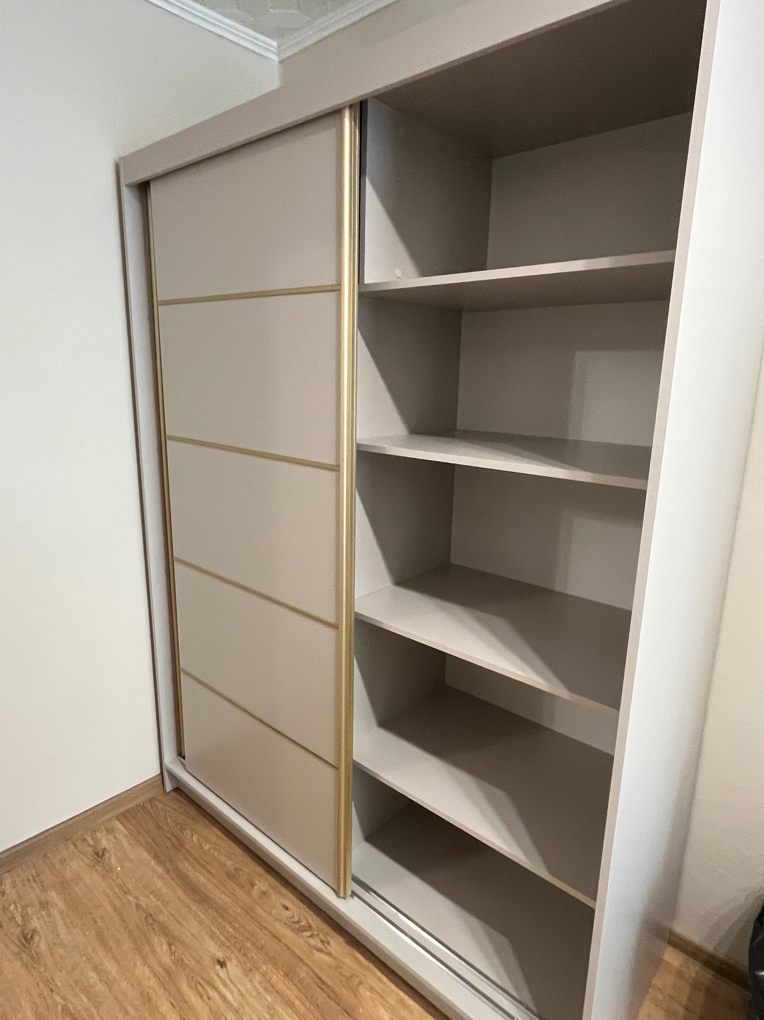 Beżowa szafa przesuwna z złotymi akcentami, widoczne półki wewnętrzne, stojąca na drewnianej podłodze. Nowoczesny design.